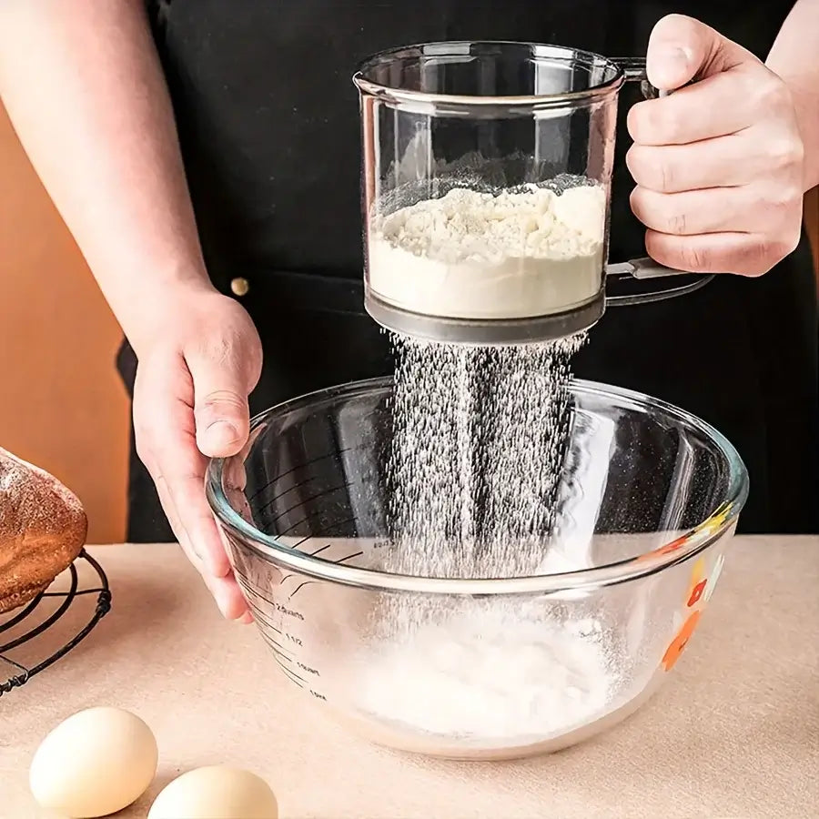 Person sifting flour into a glass bowl with eggs on a wooden surface.
