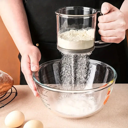 Person sifting flour into a glass bowl with eggs on a wooden surface.