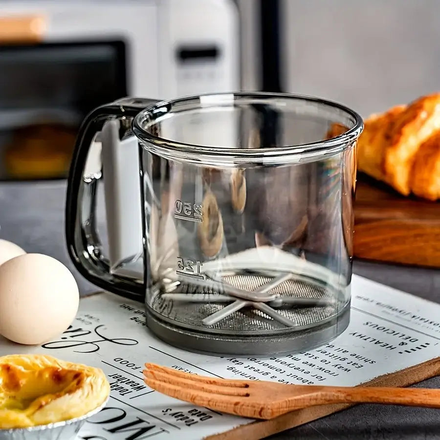 Clear glass mug with metal strainer on a newspaper, surrounded by eggs and pastries.
