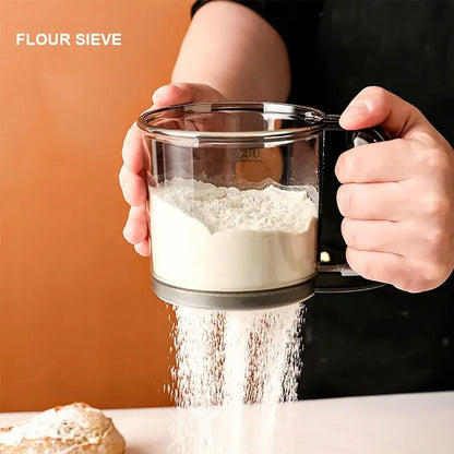Person using a flour sieve to sift flour into a glass container with a plain background.